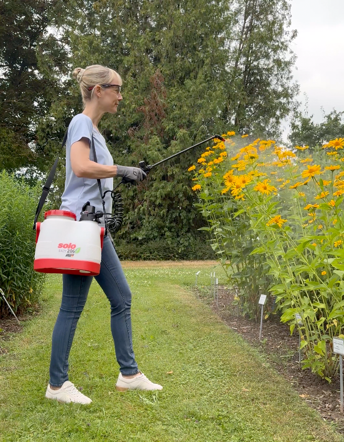 Frau mit SOLO 206 Li Akku-Sprüher sprüht gelbe Blüten in einem Garten, ausgerüstet mit Schutzhandschuhen und Schutzbrille.
