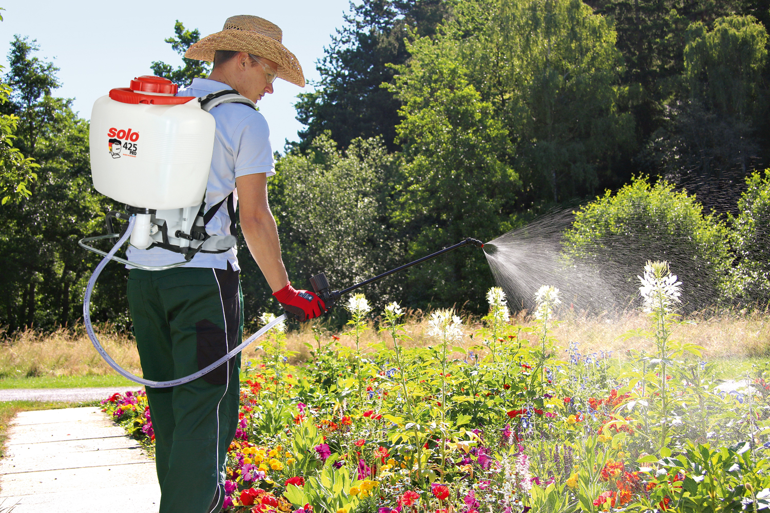 Mann mit Strohhut und SOLO 425 PRO Rückenspritze besprüht ein bunt blühendes Blumenbeet bei sonnigem Wetter.
